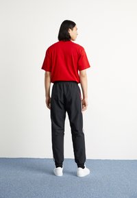 T-shirt vermelha de manga curta combinada com calças de treino pretas com cintura elástica e riscas laterais. O modelo usa ténis brancos.