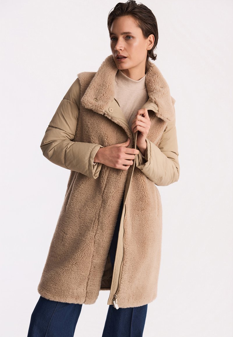 Beżowy płaszcz z owczej wełny z wyściełaną warstwą zewnętrzną i szerokim kołnierzem. Charakteryzuje się zapięciem na napy i suwak. Miękka tekstura i długość do kolan.