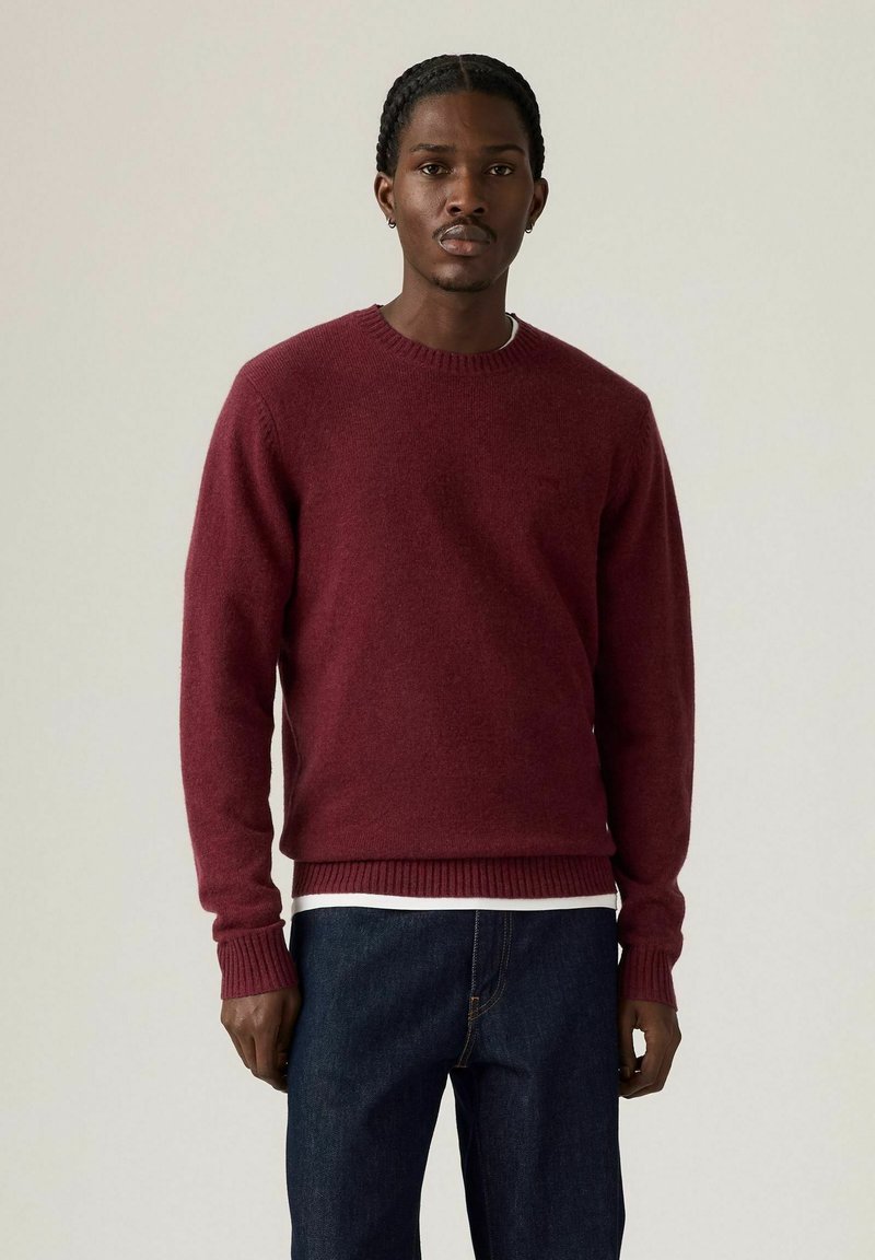Bordeaux wollen trui met een ronde hals en geribbelde manchetten en zoom, gedragen over een wit shirt, gecombineerd met donkere denim jeans.