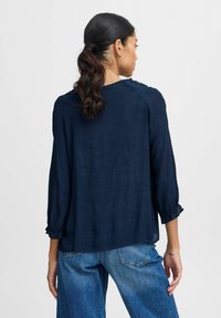 Navy blue, lightweight blouse with three-quarter sleeves, gathered cuffs, and textured fabric. Shown from the back. Paired with blue jeans.