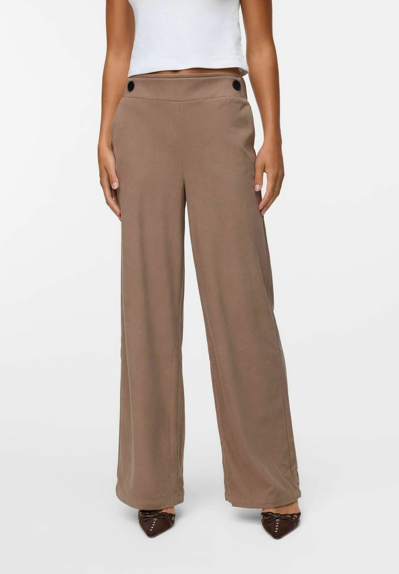 Femme portant un pantalon marron taille haute, à jambes larges, avec des boutons noirs et des chaussures pointues foncées, associé à un top court blanc.