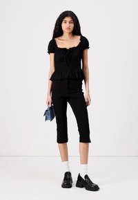 Crna peplum bluzica s nabranim izrezom i kratkim rukavima, u kombinaciji s pripijenim crnim hlačama do članka. Model drži plavu torbu i nosi crne cipele.