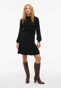 Pull en maille noir à manches longues et coupe décontractée, associé à une jupe évasée noire. Le modèle porte des bottes en cuir marron à hauteur de genou.