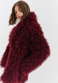 Bordó bunda z umělé kožešiny s chlupatou texturou, oversized střih, široké rukávy a vysoký límec, na neutrálním pozadí.