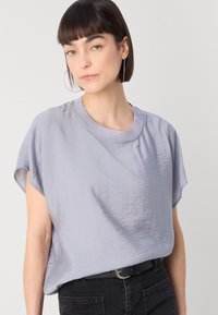 Haut gris clair à manches courtes avec une texture légèrement brillante et un col rond, associé à un jean en denim foncé. Le design présente des manches amples.