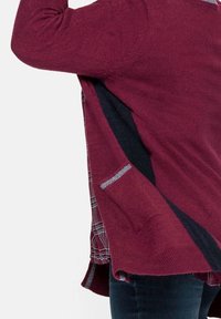 Il cardigan in maglia bordeaux presenta un design a strati con orlo a quadri, tasche laterali e dettagli neri. Texture morbida, silhouette rilassata.