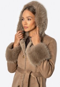 Brązowy zamszowy płaszcz z kapturem i mankietami wykończonymi futrem, z paskiem do związania i zapięciem na guziki. Miękka tekstura i ciepły design.