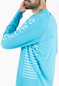 Lys blå langærmet træningsskjorte lavet af et glat, strækbart stof. Har hvid lodret tekst på den ene ærme og vandrette striber på siden.