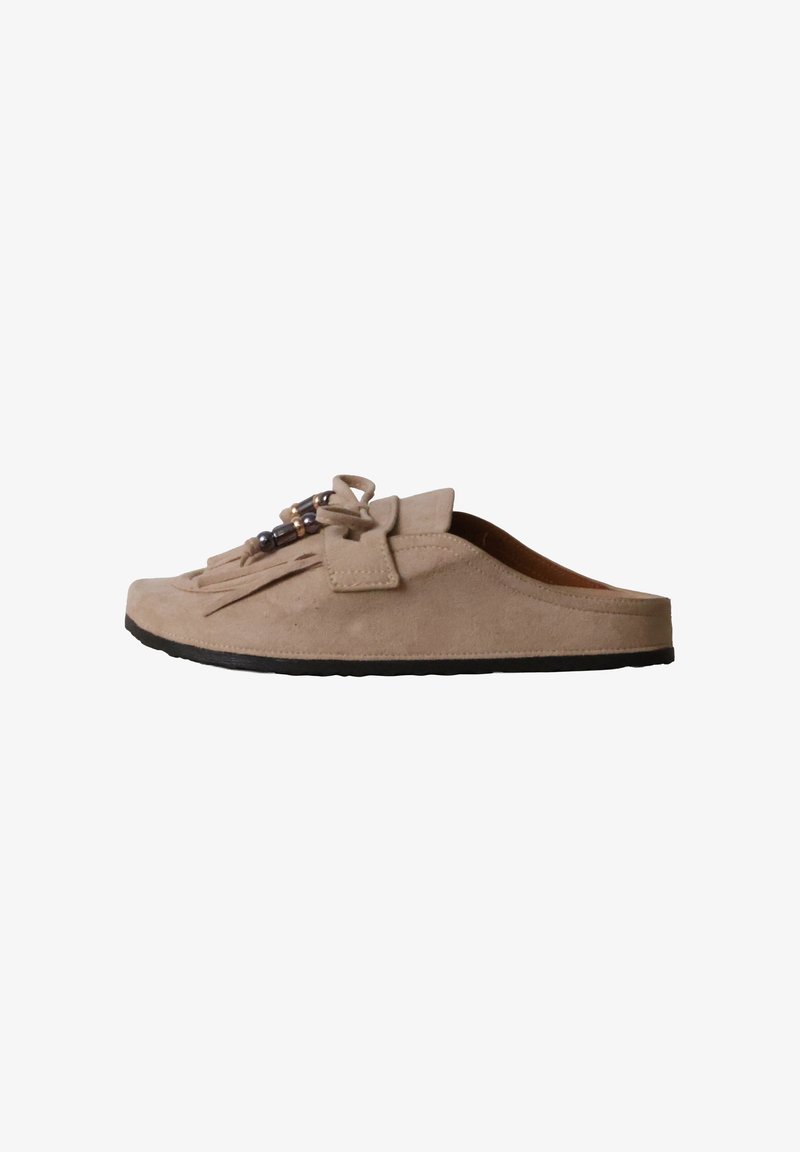 Mocassin beige en daim à enfiler avec semelle noire, pompons à l'avant, nœud et perles décoratives sur fond blanc.