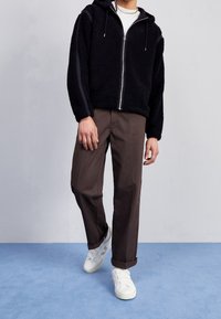 Schwarze Fleece-Zip-up-Hoodie mit Kapuze, kombiniert mit braunen geraden Hosen und weißen Sneakers. Mit kontrastierenden schwarzen seitlichen Streifen.