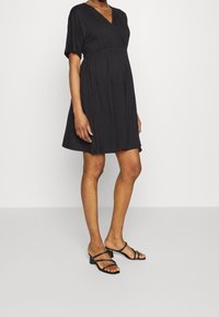 Czarna sukienka na krótki rękaw z dekoltem w serek, rozkloszowanym dołem i cinchowanym w talii. Noszona z czarnymi sandałami na paski, ukazującymi gładką strukturę i prosty design.