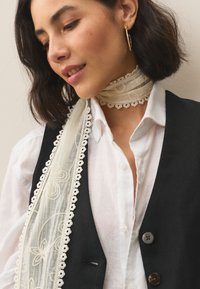 Črni telovnik čez belo srajco z ovratnikom, kombiniran z prozornim šalcem z mehkimi čipkami in cvetličnim vezenjem. Viden zlati obročkov uhani.