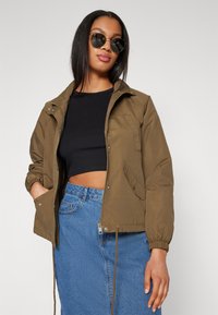 Giacca bomber khaki con zip frontale, polsini elasticizzati e orlo con cordoncino. Abbinata a un top corto nero e una gonna di jeans blu.