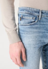 Jeans de denim azul claro com um acabamento desbotado, apresentando um logo no bolso de trás, botão metálico e detalhes de costura. Manga de um suéter bege suave visível.