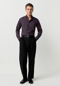 Dunkelviolettes Langarmhemd mit Tasche, schwarze Hose und schwarze Schuhe. Das Hemd hat Knöpfe und strukturierte Manschetten.