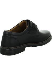 Scarpa nera in pelle con tacco basso, dotata di una cinghia in velcro e dettagli di cucitura. Texture liscia con un design a punta rotonda.
