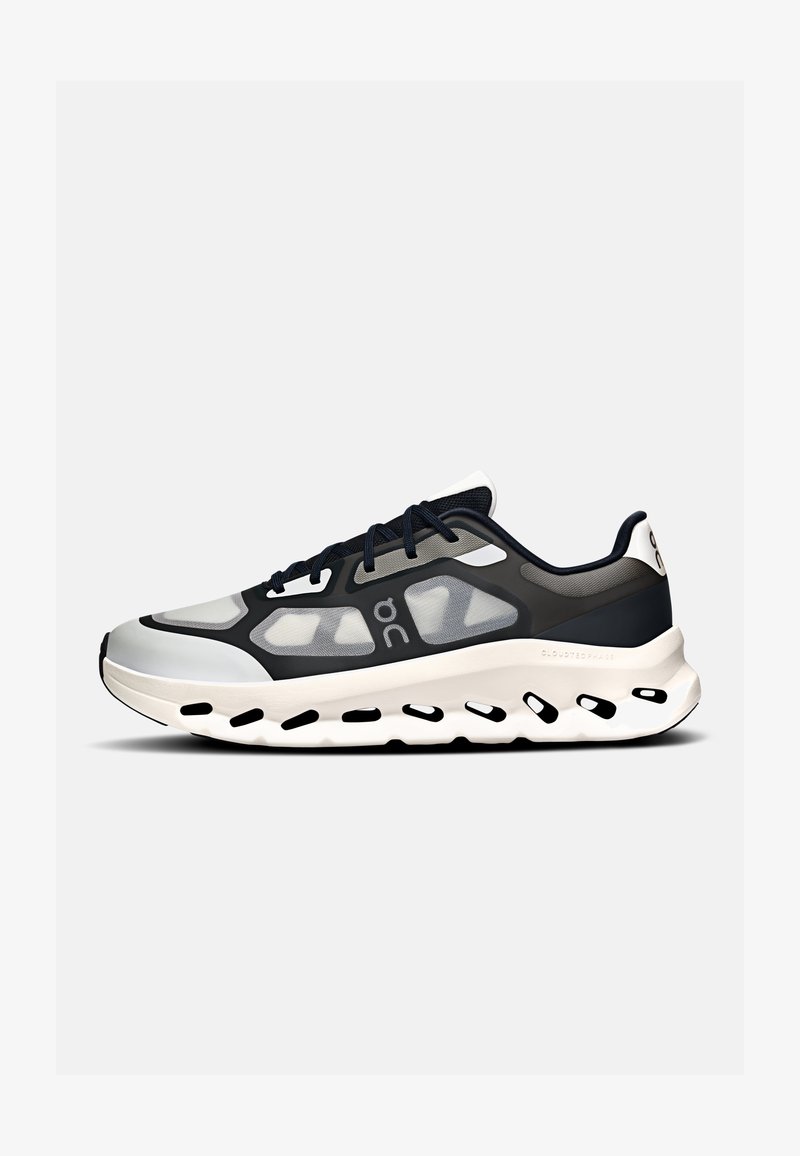 Chaussure de sport gris clair et noir avec panneaux latéraux translucides, lacets noirs, semelle blanche rembourrée avec découpes noires et logo de la marque sur le côté.