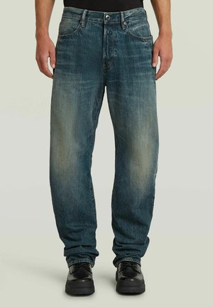 Uomo che indossa jeans blu slavati a taglio ampio con tasche anteriori e scarpe nere con lacci, in piedi contro uno sfondo semplice.