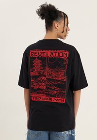 Frau mit lockigem Haar in einem Dutt, trägt ein schwarzes T-Shirt mit rotem Motiv eines Berges, einer Pagode und einer Landschaft, mit dem Text „Revelation“ und „Find Your Path“.