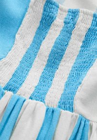 Smockitud kangas detail, millel on vahelduvad helesinise ja valge triibud, ning allosas on kogutud kortsud, mis lisavad tekstuuri ja kuju.