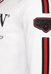 Vit bomullströja med röda och svarta randiga detaljer på ärmarna och en svart broderad "TOP GUN"-patch på ärmen.