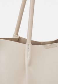 Sac cabas en cuir beige avec surface texturée et anses cousues, montrant un gros plan de la fixation de l’anse et du bord supérieur.