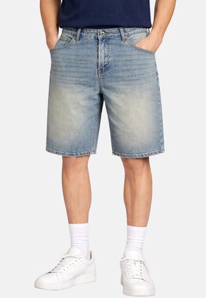 Uomo con indosso pantaloncini di jeans azzurri sbiaditi, calzini bianchi, scarpe da ginnastica bianche e una camicia blu scuro con le mani in tasca.