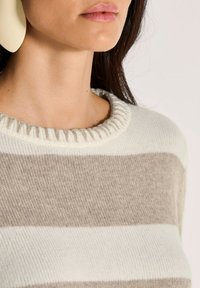 Frau trägt einen beigen und weißen gestreiften Strickpullover mit Rippenhalsausschnitt, zeigt Hals und Kinn vor einem schlichten Hintergrund.