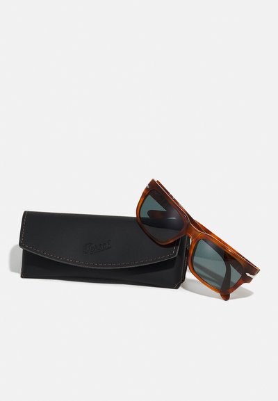 Occhiali da sole tortoiseshell marroni con lenti scure, esposti accanto a un astuccio in pelle nera con logo impresso e cuciture a contrasto.