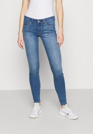 Personne portant un jean skinny bleu, un débardeur blanc et des baskets blanches, debout devant un fond clair uni.