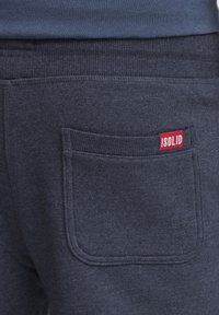 Navyblå sweatshirtbyxor har en bakficka med ribbad kant och en liten röd etikett med texten "!SOLID." Mjuk textur, avslappnad design.
