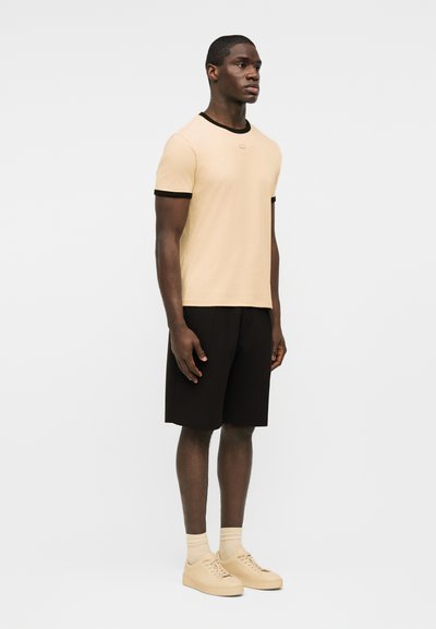 Άνδρας όρθιος που φοράει μπεζ μπλουζάκι με μαύρη επένδυση, μαύρο σορτς, μπεζ κάλτσες και μπεζ αθλητικά παπούτσια, πάνω σε έναν απλό φωτεινό φόντο.