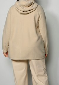 Chaqueta de forro polar beige con capucha y mangas largas, con un tejido texturizado y un corte holgado, combinada con pantalones de pierna ancha beige claro.