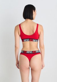 Ensemble de lingerie rouge en matériau extensible. Le soutien-gorge a des bretelles larges et une bande noire avec le logo "DIESEL". Le slip présente un design de ceinture similaire.