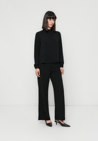 Zwarte lange mouwen top en wijde broek van gladde stof, gecombineerd met zwarte puntige hakken, op een effen achtergrond.