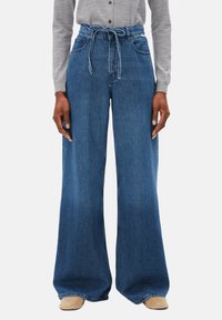 Jean en denim à jambes larges de couleur bleu moyen, avec une taille haute et un lien à nouer, accompagnés de poches latérales. Le tissu a une texture lisse.