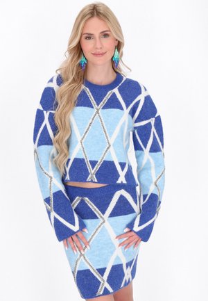Lyshåret kvinde iført blå og lyseblå diamantmønstret sweater og matchende nederdel, med langt bølget hår og farverige lagdelte øreringe.