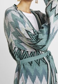 Kvinna i en mönstrad stickad cardigan i teal och vitt med ett bälte knytet i midjan, tillsammans med en vit skjorta och guld geometriska örhängen.