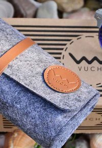 Šedý měkký sáček s béžovým koženým popruhem a kulatou nášivkou s logem. Vyznačuje se zjednodušeným designem a jemnou texturou, umístěný na texturovaném pozadí.