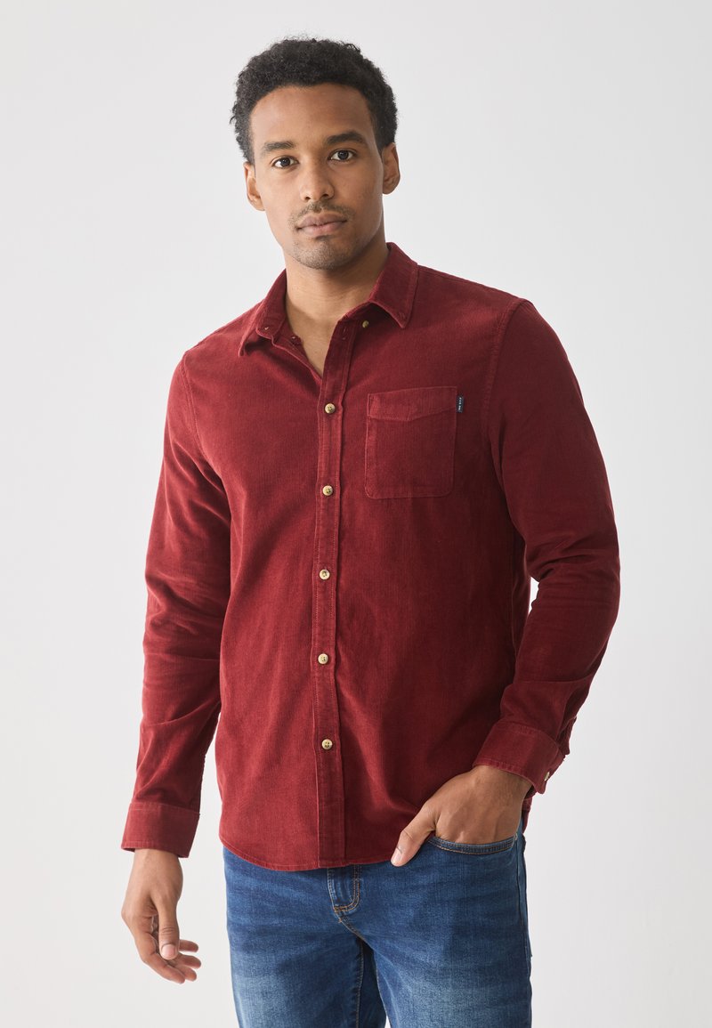 Camisa de pana roja con botones y un bolsillo en el pecho, botones marrones y mangas largas, combinada con jeans de mezclilla azules.