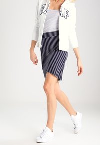 Mujer caminando, vestida con un hoodie blanco de GAP, una camiseta de tirantes gris, una falda a rayas azul marino y blanca, y zapatillas blancas sobre un fondo claro.