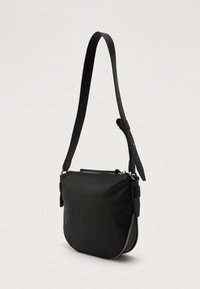 Μαύρη δερμάτινη τσάντα ώμου με ρυθμιζόμενο λουράκι, κλείσιμο με φερμουάρ και διακριτό ανάγλυφο λογότυπο μπροστά, τοποθετημένη σε απλό φόντο.