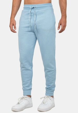 Mężczyzna ubrany w jasnoniebieskie spodnie dresowe ze ściągaczem w talii i białe sneakersy, stojący na jednolitym tle.