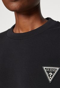 T-shirt in cotone nero con scollo a girocollo e un patch a forma di triangolo che presenta il testo "GUESS" e un punto interrogativo sul lato sinistro del petto.