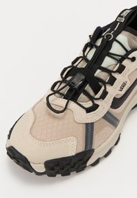 Beige mesh- och mockaskor med svarta accenter, elastiska snören och mönstrad sula; har en förstärkt tå och vadderad krage för komfort.