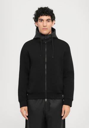 Svart zip-up hettegenser med teksturert svart hette, snor og sidelommer; har en avslappet passform og ribbede ermer.