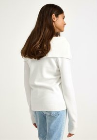 Maglione bianco con collo e polsini a coste, caratterizzato da un design aderente e una texture morbida, abbinato a jeans azzurri.