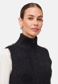 Žena v čiernej chlpaté bezrukávovej zipsovej veste a bielej dlhoprstej košeli, s zlatými kruhovými náušnicami na pozadí s jednoduchým vzorom.
