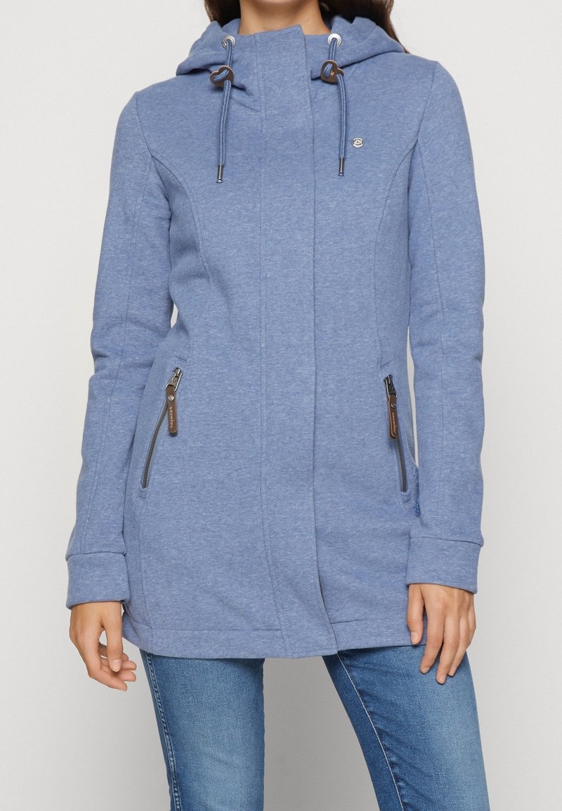Femme portant une veste à capuche bleue avec cordons de serrage et poches latérales zippées, associée à un jean bleu, visible des épaules aux hanches.