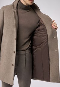Cappotto di lana color marrone chiaro, con foderatura liscia, due bottoni e vestibilità su misura, indossato sopra un dolcevita e pantaloni coordinati.